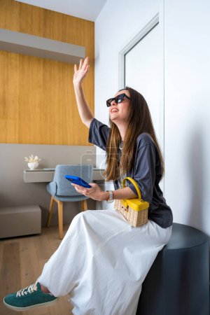 Une femme souriante portant des lunettes de soleil est assise à l'intérieur, portant une jupe blanche en ballon, des baskets en sarcelle et tenant un sac à main en osier avec une poignée jaune. Elle agite la main tout en tenant un smartphone bleu.