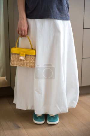 Eine Frau steht drinnen im weißen Ballon-Midi-Rock und teal-Turnschuhen und hält eine stylische Weidenhandtasche mit gelbem Griff in der Hand. Der moderne feminine Casual Look.