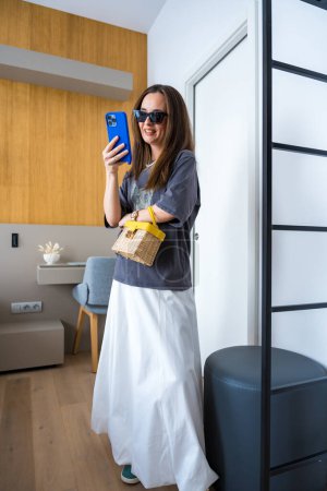Femme souriante aux lunettes de soleil oeil de chat debout à l'intérieur, portant une jupe en ballon blanc, baskets turquoise, et tenant un sac à main en osier avec une poignée jaune. Elle tient un smartphone bleu et le regarde.