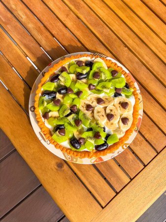 A delicious homemade open pie topped with sour cream, kiwi, banana, and grapes, sprinkled with chocolate hearts. The rustic wooden table adds a cozy, homey feel to this delightful dessert.