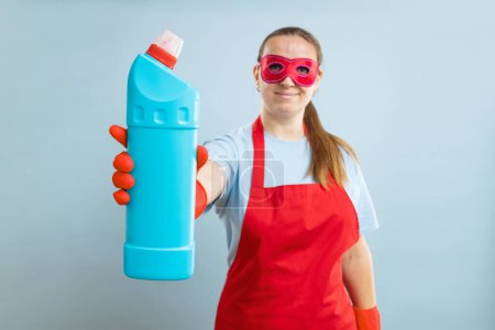 Téléchargez les photos : Jeune femme en masque rouge, gants en caoutchouc et tablier tenant bouteille de détergent sur fond bleu. Détergent publicitaire - en image libre de droit