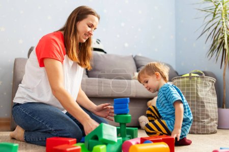 Cute little kid and child development specialist attractive young woman playing together with colorful blocks, sitting on the floor.