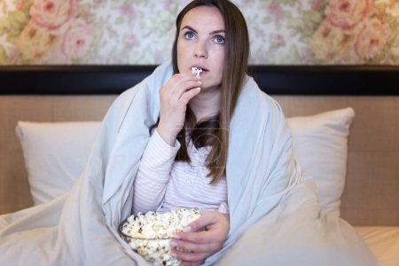 Young woman is watching projector, TV, movies and eating popcorn in the bed at home