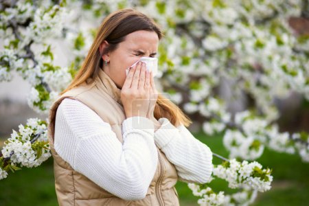 Téléchargez les photos : Femme éternuante souffrant d'allergie saisonnière au printemps avec un essuie-nez parmi les arbres à fleurs dans le parc. Concept d'allergies printanières - en image libre de droit