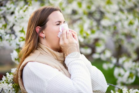 Téléchargez les photos : Femme éternuante souffrant d'allergie saisonnière au printemps avec un essuie-nez parmi les arbres à fleurs dans le parc. Concept d'allergies printanières - en image libre de droit