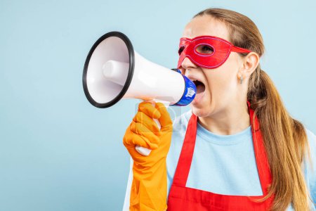 Photo for Cleaning woman in red hero mask, gloves and apron with megaphone on blue background. Announcing information of cleaning services - Royalty Free Image