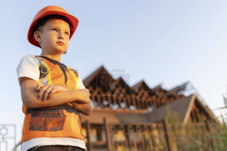 A young child stands with arms crossed wearing a safety vest and hard hat at a residential construction site during sunset.