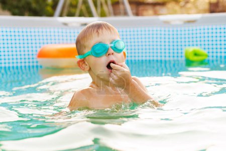 Fröhlicher kleiner Junge mit Schwimmbrille steigt in einem Pool aus dem Wasser, den Mund mitten im Gespräch oder Lachen aufgerissen und genießt ein erfrischendes Bad.