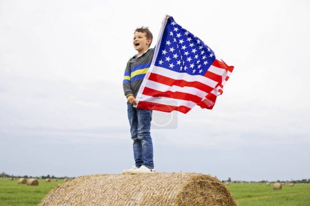 Joyful young boy standing on a hay bale, triumphantly waving the American flag with a clear sky in the background.