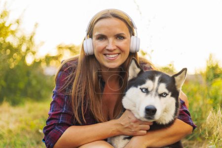 In a golden hour glow, a smiling woman warmly embraces her loyal Husky dog while enjoying a serene outdoor setting.
