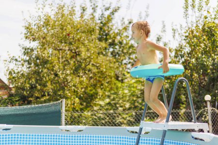 Foto de Muchacho emocionado con un flotador de tabla de surf azul preparándose para saltar a una piscina sobre el suelo en un día soleado de verano, rodeado de vegetación. - Imagen libre de derechos