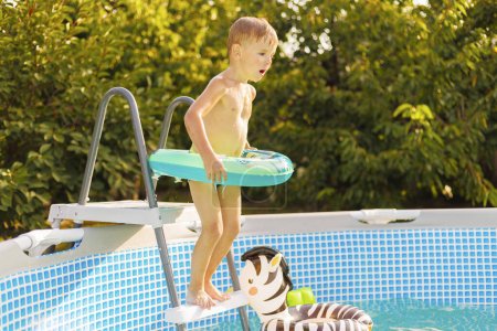 Ein kleiner Junge mit Schwimmring steht an einer Schwimmbadleiter, aufgeregt und bereit, an einem sonnigen Tag ins Wasser zu springen.