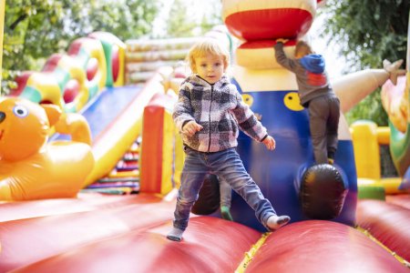 Téléchargez les photos : Jeune enfant jouant sur un château plein d'entrain gonflable vibrant dans une aire de jeux extérieure entourée de verdure. - en image libre de droit