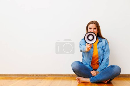 Casual woman holding a megaphone, sitting cross-legged on a wooden floor against a white wall