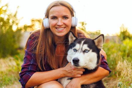 In a golden hour glow, a smiling woman warmly embraces her loyal Husky dog while enjoying a serene outdoor setting.