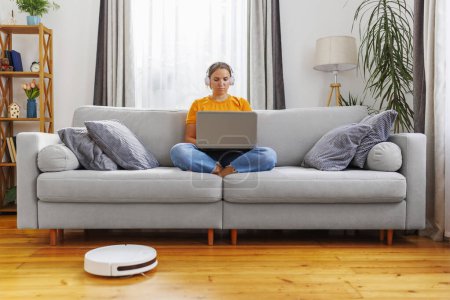 A focused woman sits cross-legged on a sofa working on a laptop with headphones, embodying the remote work lifestyle.