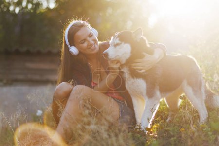 Eine fröhliche junge Frau mit Kopfhörern umarmt ihren Siberian Husky in einer sonnenbeschienenen natürlichen Umgebung und teilt einen Moment der Entspannung.