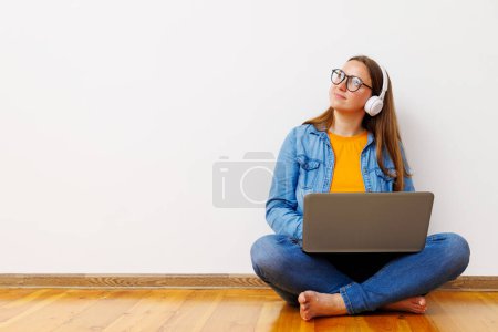 Eine entspannte junge Frau in Freizeitkleidung sitzt im Schneidersitz auf dem Boden, hört Musik mit Kopfhörern und benutzt einen Laptop.