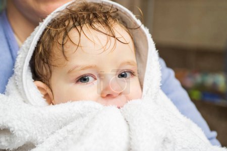 A fresh-faced toddler with wet hair is snuggled in a white fluffy towel, held by a parent after a warm bath.