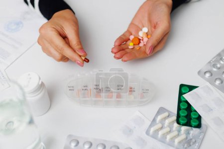 Close-up of hands sorting daily medication into a pill organizer, with prescriptions and water on the table.
