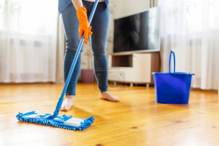Unidentifiable person in casual clothing mopping a wooden floor with a blue mop and bucket in a bright room.