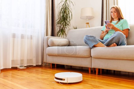 Eine junge Frau lümmelt auf einer Couch und bedient ihr Smartphone, während ein Staubsaugerroboter den Laubholzboden reinigt.