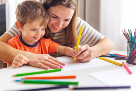 A young boy grins broadly as his mother assists him in drawing with pencils, enjoying a creative afternoon together.