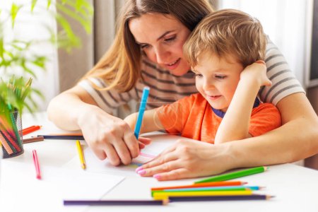 A young mother is engaging with her little son in a fun drawing activity using colorful pencils on a bright day.