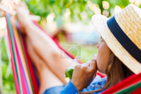 Una mujer disfruta de un día de verano perezoso, bebiendo una bebida fresca mientras se relaja en una hamaca, sombreada por una vegetación de jardines.