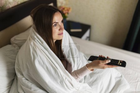 Entspannte Frau, die mit einer Decke um sich gewickelt im Bett sitzt, eine Fernbedienung in der Hand hält und fernsieht.