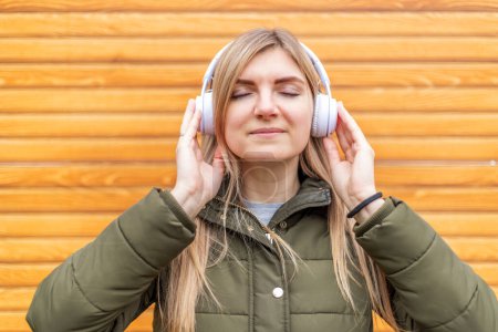 Eine zufriedene junge Frau mit geschlossenen Augen genießt Musik auf weißen Kopfhörern vor einem hölzernen Hintergrund.