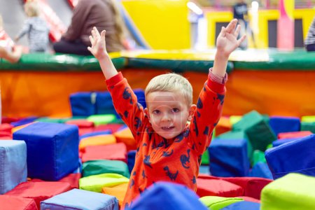 A joyful toddler boy raises his hands in a vibrant soft play area filled with colorful foam blocks.