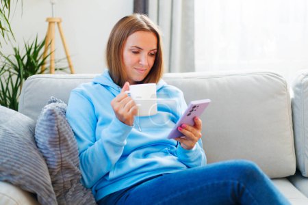 Mujer relajada en una sudadera azul bebiendo café mientras navega en su teléfono inteligente, cómodamente sentada en un sofá.