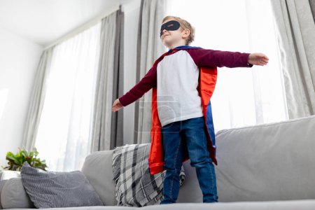 Niño pequeño imagina ser un superhéroe, de pie con confianza en una sala de estar con una capa.