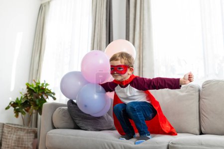 Boy in superhero costume with red cape and mask playing with balloons. Home indoor leisure activity. Childhood imagination and play concept for design and print