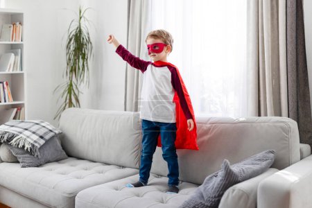 Téléchargez les photos : Enfant en costume de super héros avec cape rouge et masque debout sur un canapé. Jeu imaginaire et concept d'enfance. Intérieur de la maison prise de vue avec lumière naturelle et espace pour le texte - en image libre de droit