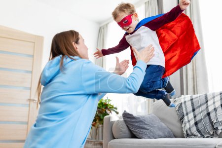 Téléchargez les photos : Mère jouant avec l'enfant en costume de super-héros. Concept d'activités de loisirs en intérieur. Conception pour la parentalité, plaisir en famille - en image libre de droit