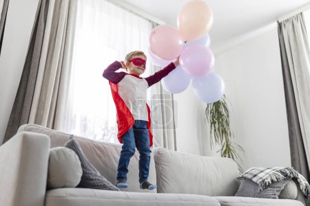 A young child dressed as a superhero stands proudly on a sofa, joyfully playing with colorful balloons.