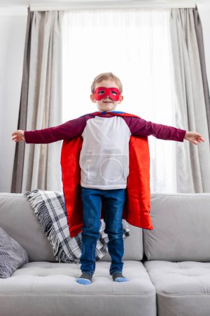 Téléchargez les photos : Enfant en tenue de super héros avec cape et masque, intérieur de la maison. Photographie de style de vie candide pour affiche et bannière. - en image libre de droit