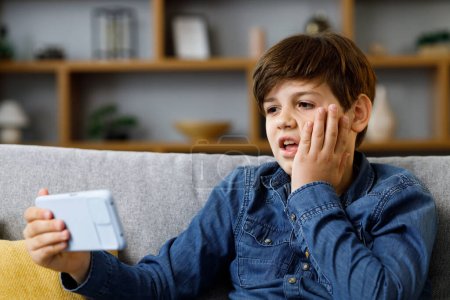 Téléchargez les photos : Young boy reacts emotionally to what is happening on the smartphone screen. Teenager spending time at home with digital gadget. Adolescence and puberty. - en image libre de droit