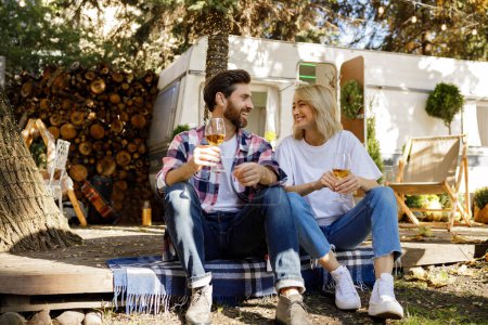 Téléchargez les photos : Un beau couple est assis près de la caravane de voyage, riant en s'amusant, profitant de leurs vacances. Les amoureux profitent d'une journée ensoleillée en dehors de la ville, boire du vin - en image libre de droit