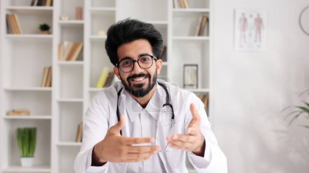 Téléchargez les photos : Clinicien indien amical intelligent en manteau blanc utilisant un ordinateur portable pour la consultation à distance des patients dans le bureau de l'hôpital. Télémédecine. Rendez-vous en ligne avec un médecin. Vue caméra - en image libre de droit
