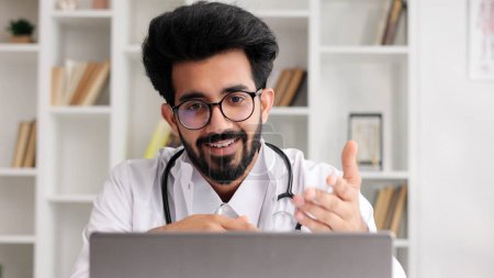 Téléchargez les photos : Clinicien indien amical intelligent en manteau blanc utilisant un ordinateur portable pour la consultation à distance des patients dans le bureau de l'hôpital. Télémédecine. Rendez-vous en ligne avec un médecin - en image libre de droit