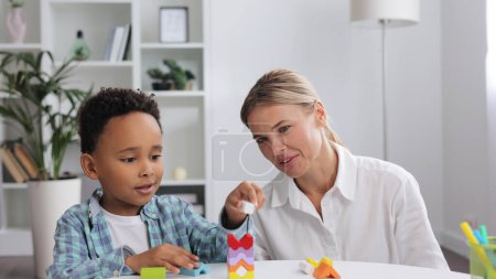 Photo for African American child curly little boy and child development specialist blonde woman playing with colorful wooden bricks sitting at table, making pyramid together. Exercises for children with autism - Royalty Free Image
