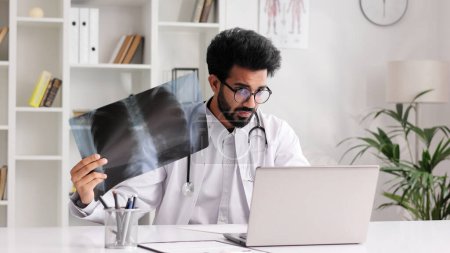 Téléchargez les photos : Smart amical homme indien clinicien en manteau blanc en utilisant un ordinateur portable pour la consultation à distance des patients dans le bureau de l'hôpital en regardant les rayons X. Télémédecine. Rendez-vous en ligne avec un médecin - en image libre de droit