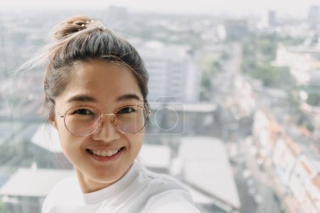 Foto de Asiática tailandesa mujer usar ojos gafas, tomando selfie con feliz sonrisa con vista a la ciudad en apartamento. - Imagen libre de derechos