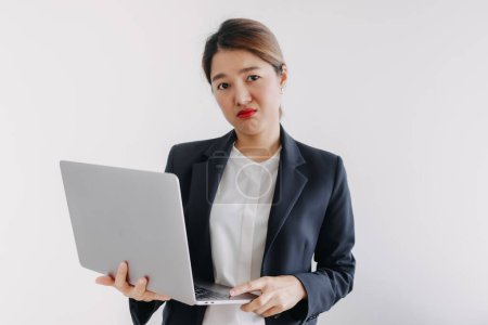 Photo for Unhappy asian Thai woman wear blazer suit, unsatisfied looking at camera with sad face, dislike holding laptop computer, standing over white background wall. - Royalty Free Image