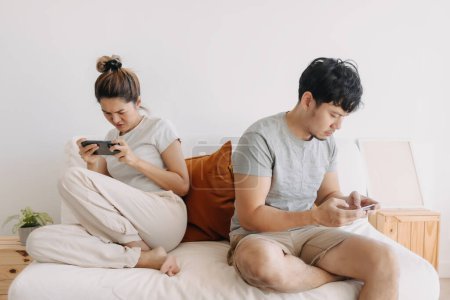 Photo for Asian Thai couple sitting back to back on sofa at home, Both man and woman using mobile phone, playing game online, ignoring living in apartment room. - Royalty Free Image