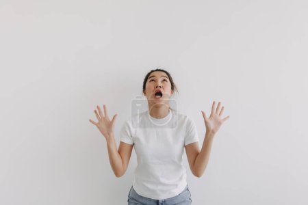Photo for Funny shocked surprised face of asian Thai woman spreading hands gesture and looking above at empty space, standing wow over white background wall. - Royalty Free Image