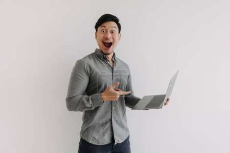 Foto de Hombre asiático con barba llevar camisa gris, wow cara sorprendida usando y sosteniendo el ordenador portátil, señalando el dedo en el cuaderno, mirando a la cámara de pie aislado sobre la pared de fondo blanco. - Imagen libre de derechos